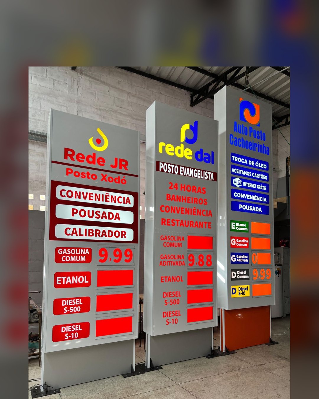 Três totens de placa de preço iluminados, das redes Rede JR, Rede Dal e Auto Posto Cachoeirinha, prontos no galpão da Tech